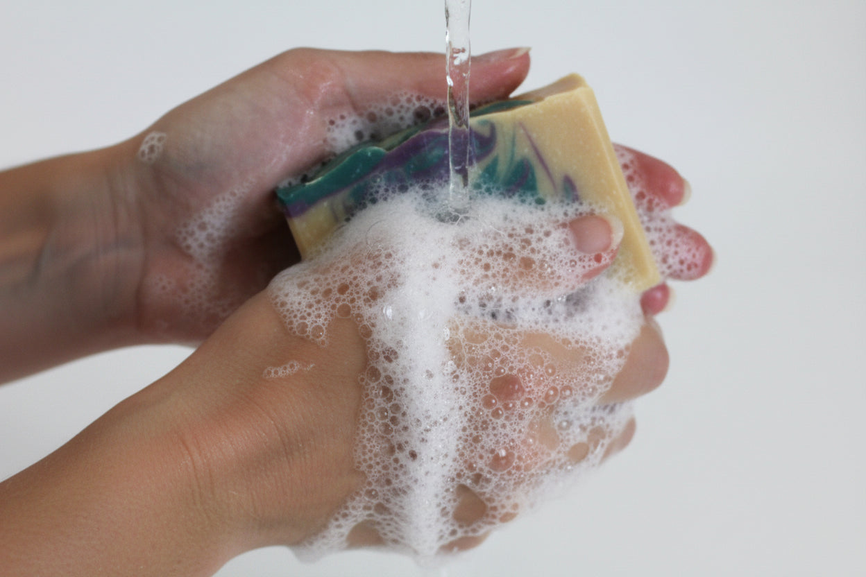 Hand holding a bar of soap with purple and green swirls on a white background being used to wash hands. There is a stream of water and bubbles.

