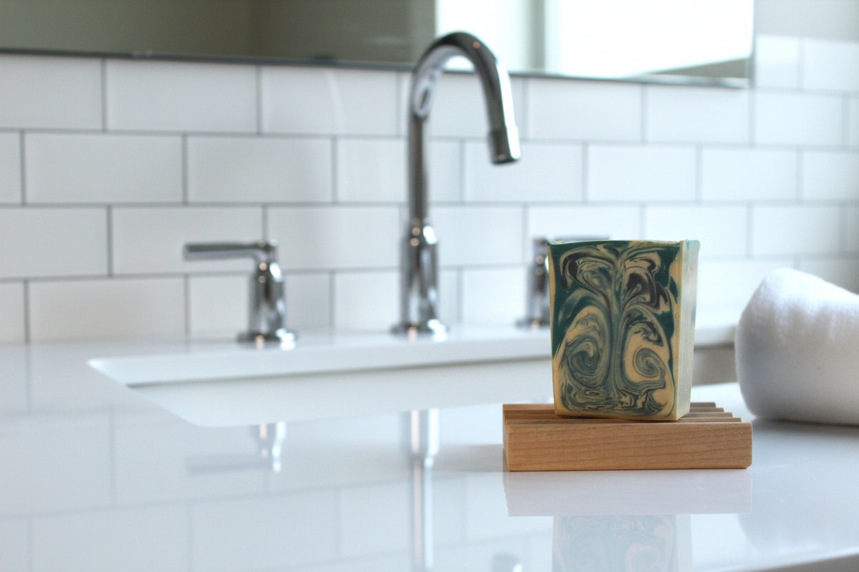 Bar of soap on a wooden block in a bathroom setting with a sink and mirror.