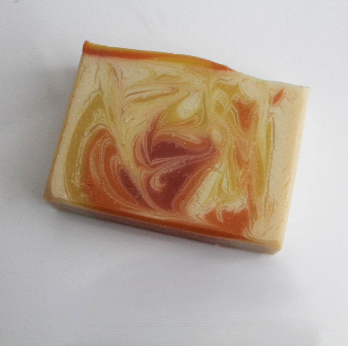 A rectangular bar of handmade soap with a marbled swirl pattern of orange, yellow, and cream, photographed on a plain white background.