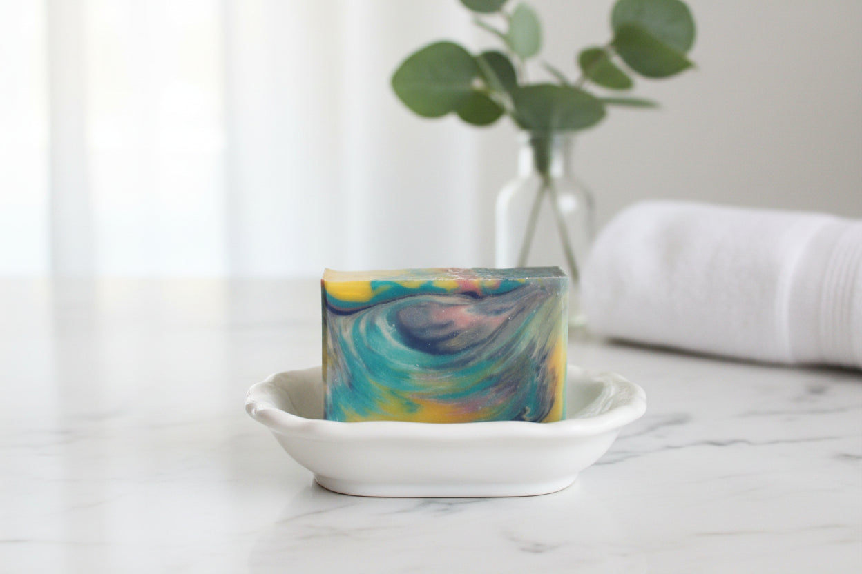 A rectangular bar of soap with a marbled swirl pattern in teal, blue, yellow, and purple, resting on a small white ceramic dish on a marble countertop. A blurred glass vase with green leaves and a rolled white towel are in the background.